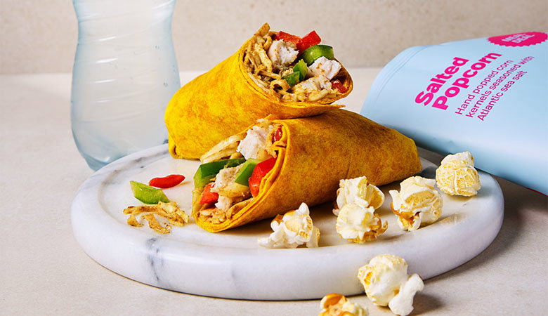 Two Katsu curry wraps filled with chicken, peppers, and onion, accompanied by popcorn and a water bottle on a marble plate.